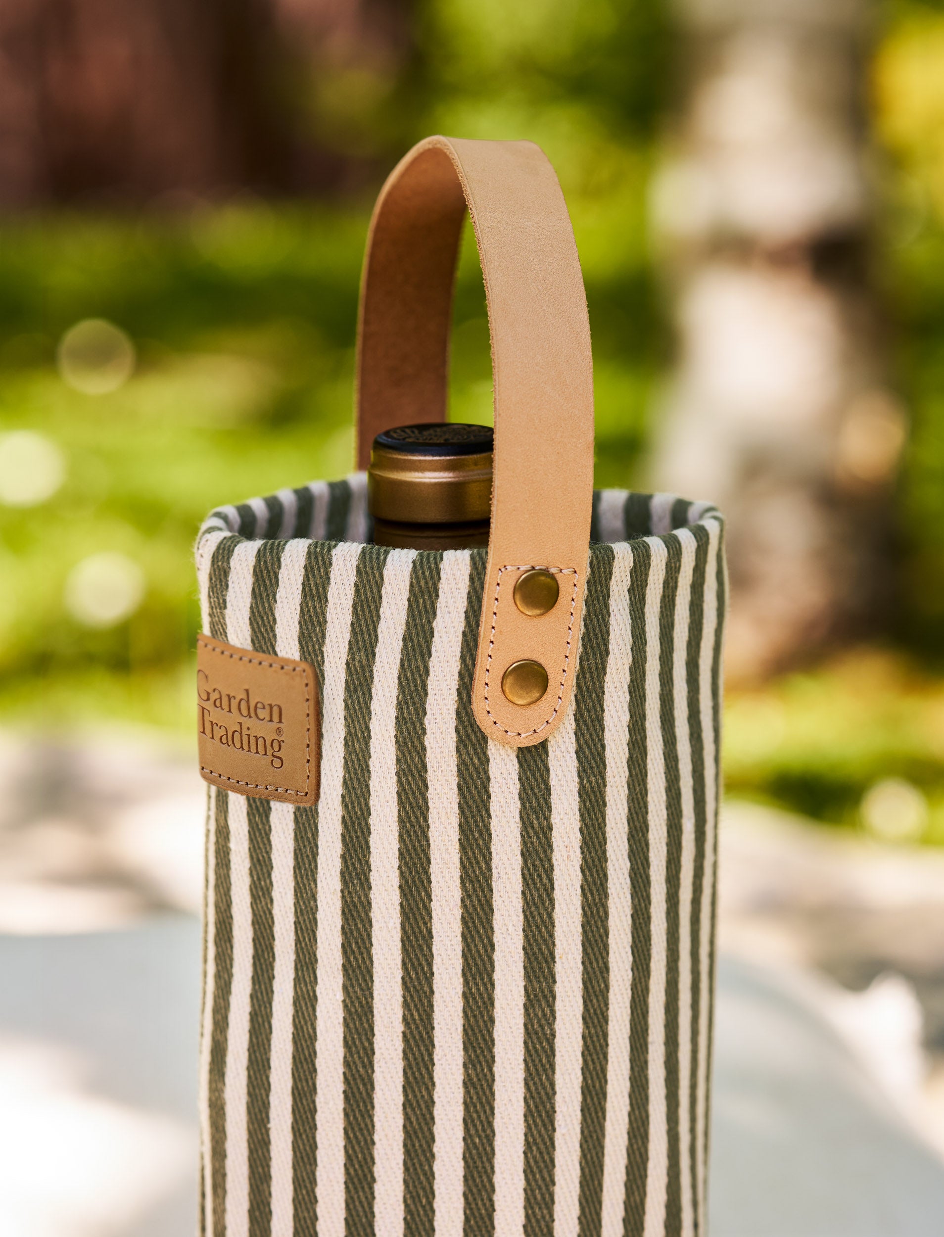 Striped wine bottle bag with leather handle and 'Garden Trading' label against a blurred green outdoor background.