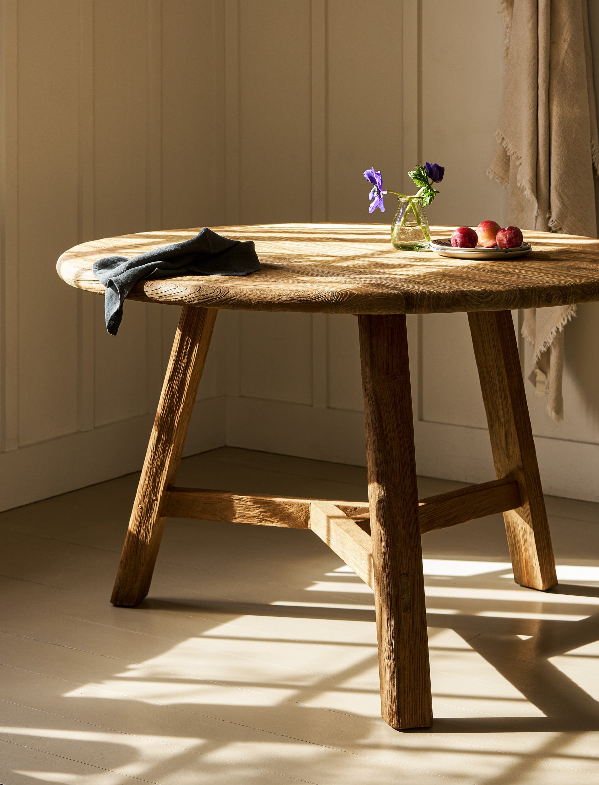 Wooden dining table with a vase of flowers and apples in a room with sunlight streaming through a window.