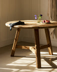 Wooden dining table with a vase of flowers and apples in a room with sunlight streaming through a window.