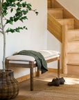Wooden bench seating with cushions and green blanket next to a staircase, with a plant and shoes on the floor.