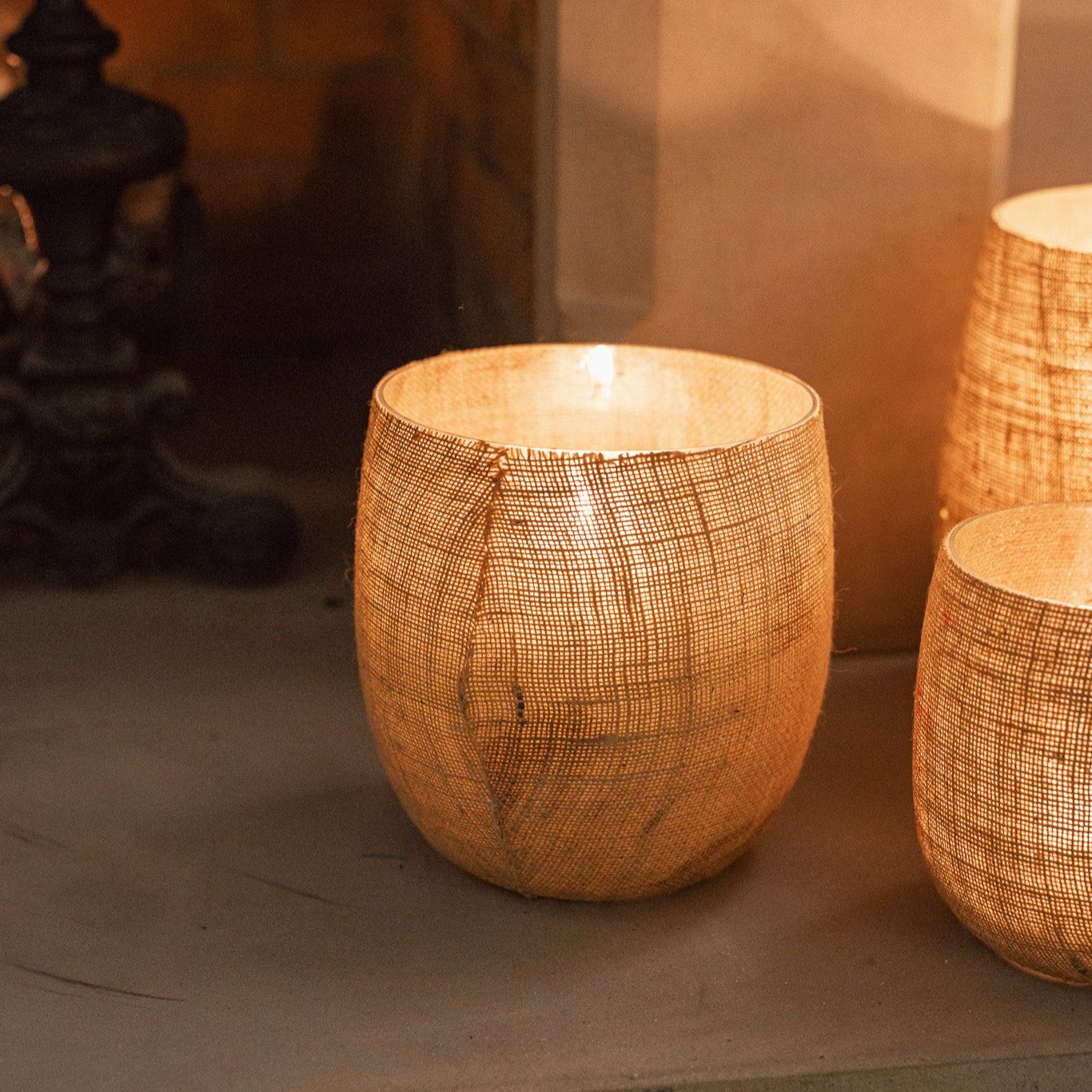 Set of three Jute Hurricane Lantern on stone fire place with lit candles inside.