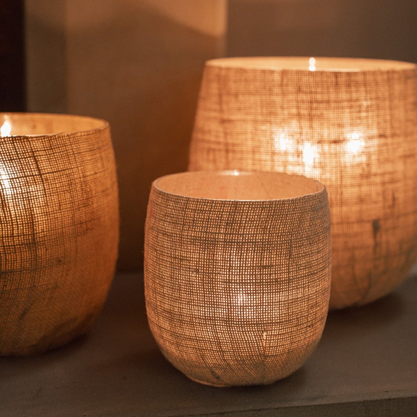 Set of three Jute Hurricane Lantern on stone fire place with lit candles inside.