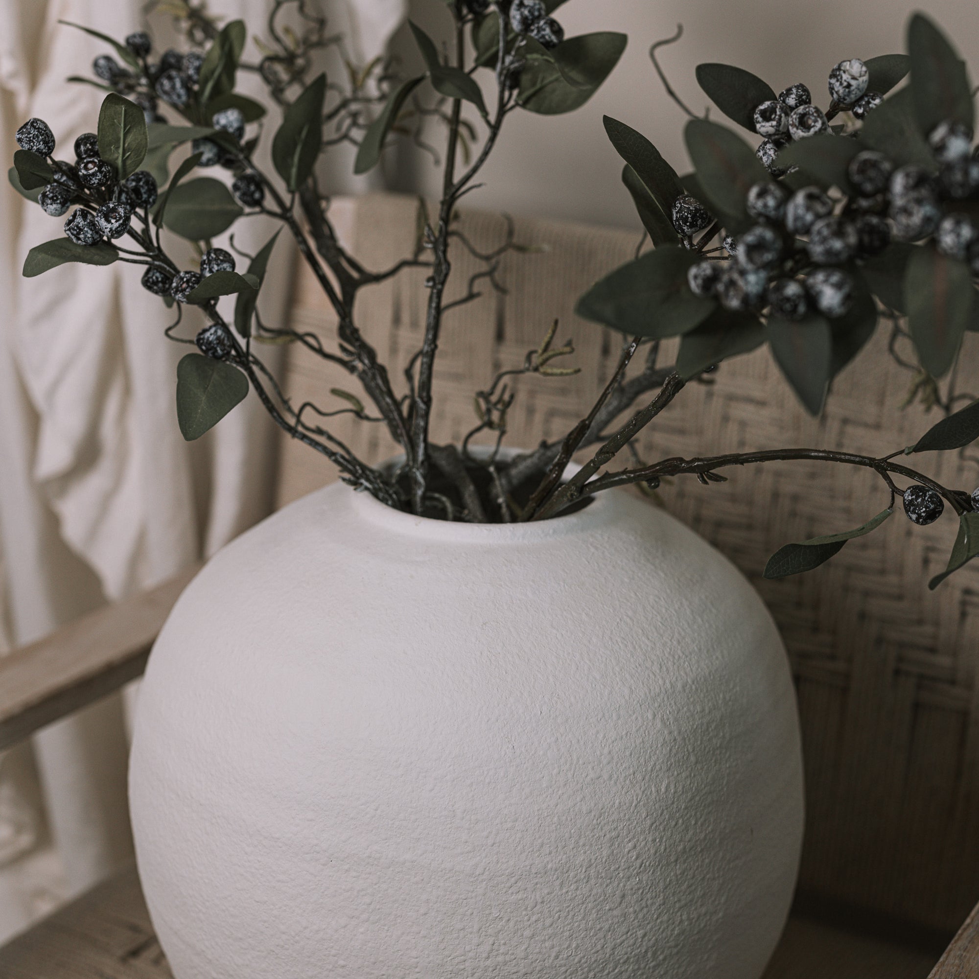Ceramic round white textured vase with berry sprays and branches on a woven armchair.
