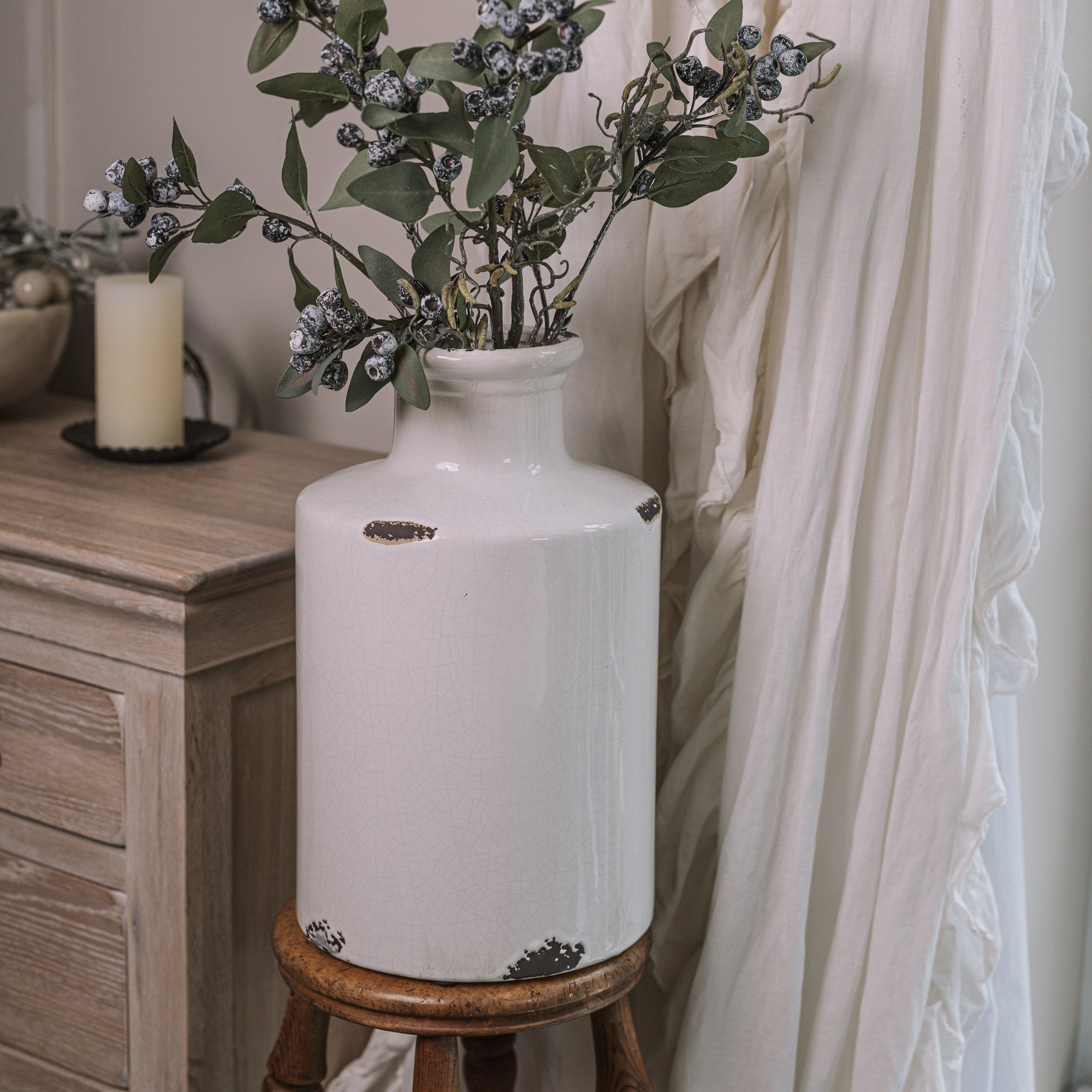 White distressed bottle vase with berry spray branches on a wooden stool.