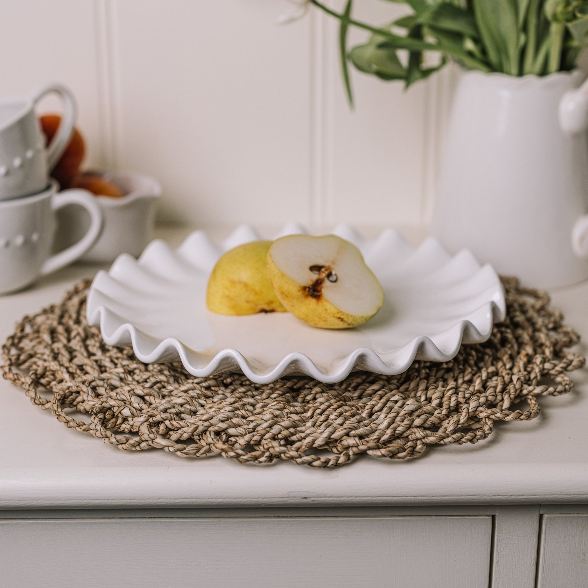 White ceramic cake plate with ruffled edges with a sliced pear on a rattan placemat.