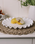 White ceramic cake plate with ruffled edges with a sliced pear on a rattan placemat.