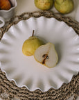 White ceramic cake plate with ruffled edges with a sliced pear on a rattan placemat.