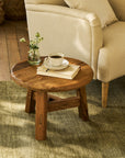 Round wooden coffee table with a cup of coffee, book, and vase of flowers next to a beige armchair.