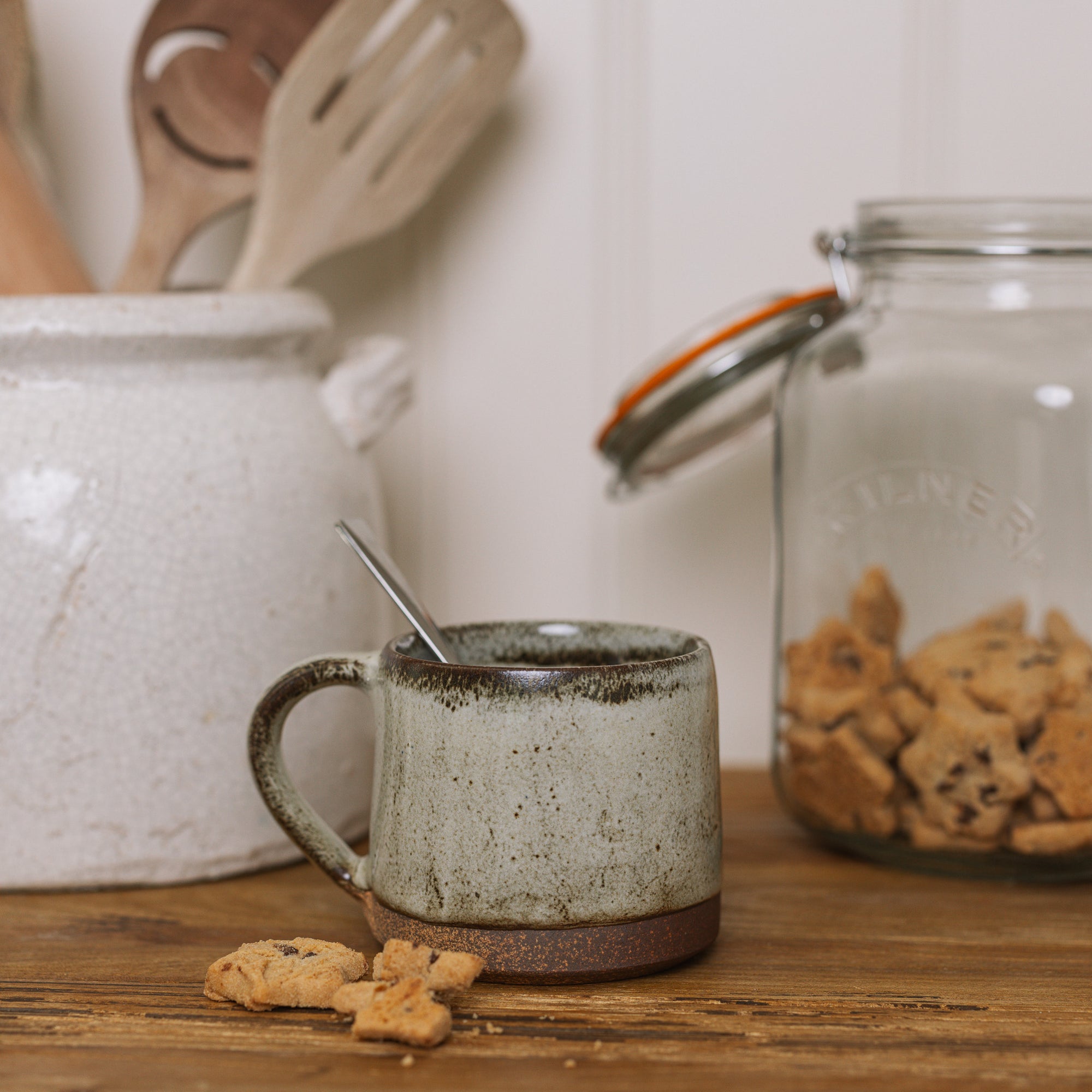 A brown speckled stoneware mug on wooden board with crumbled biscuit.