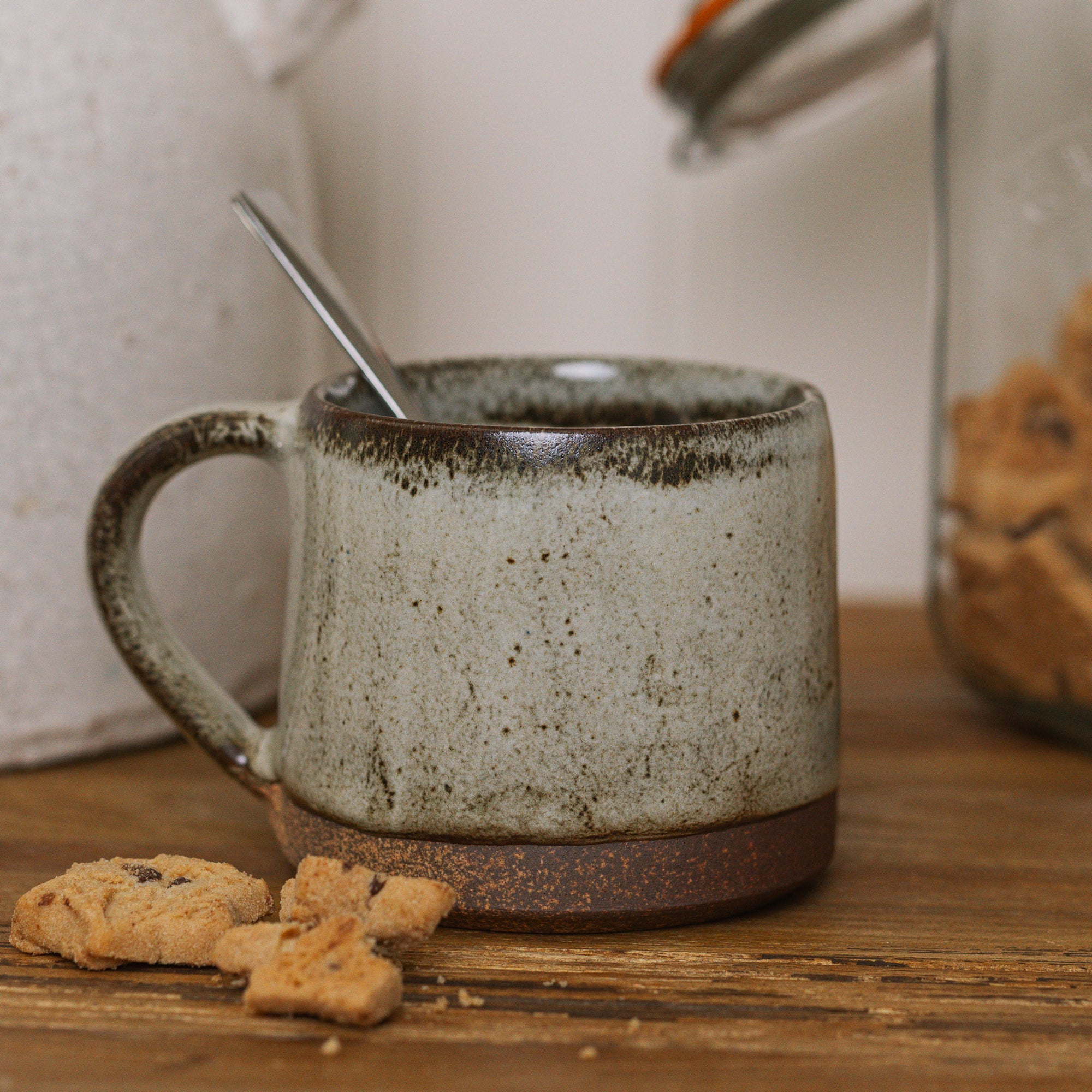 A brown speckled stoneware mug on wooden board with crumbled biscuit.