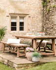 Large pine dining table outdoors with benches, plant pots and tableware.