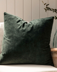 Square velvet cotton green cushion on rattan armchair with plant in vase behind.