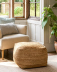 Square jute pouf in front of a cream armchair.