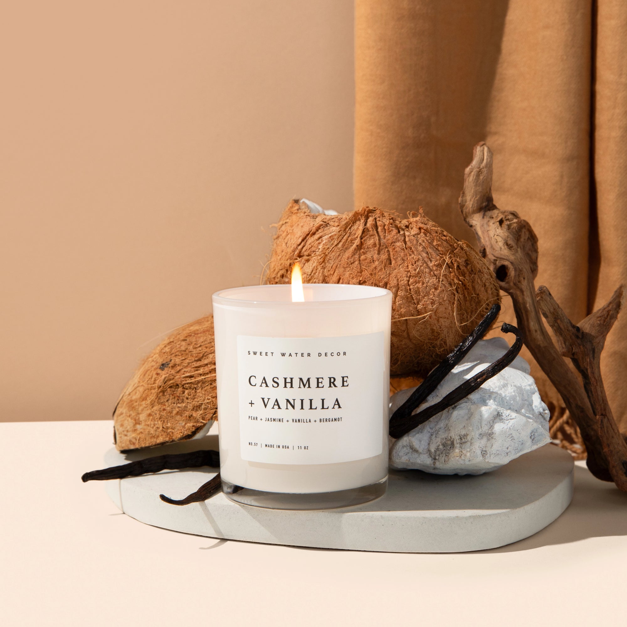 Cashmere and vanilla soy candle in white jar, lit in front of some coconuts and vanilla beans.