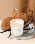 Cashmere and vanilla soy candle in white jar from above, lit in front of some coconuts and vanilla beans.