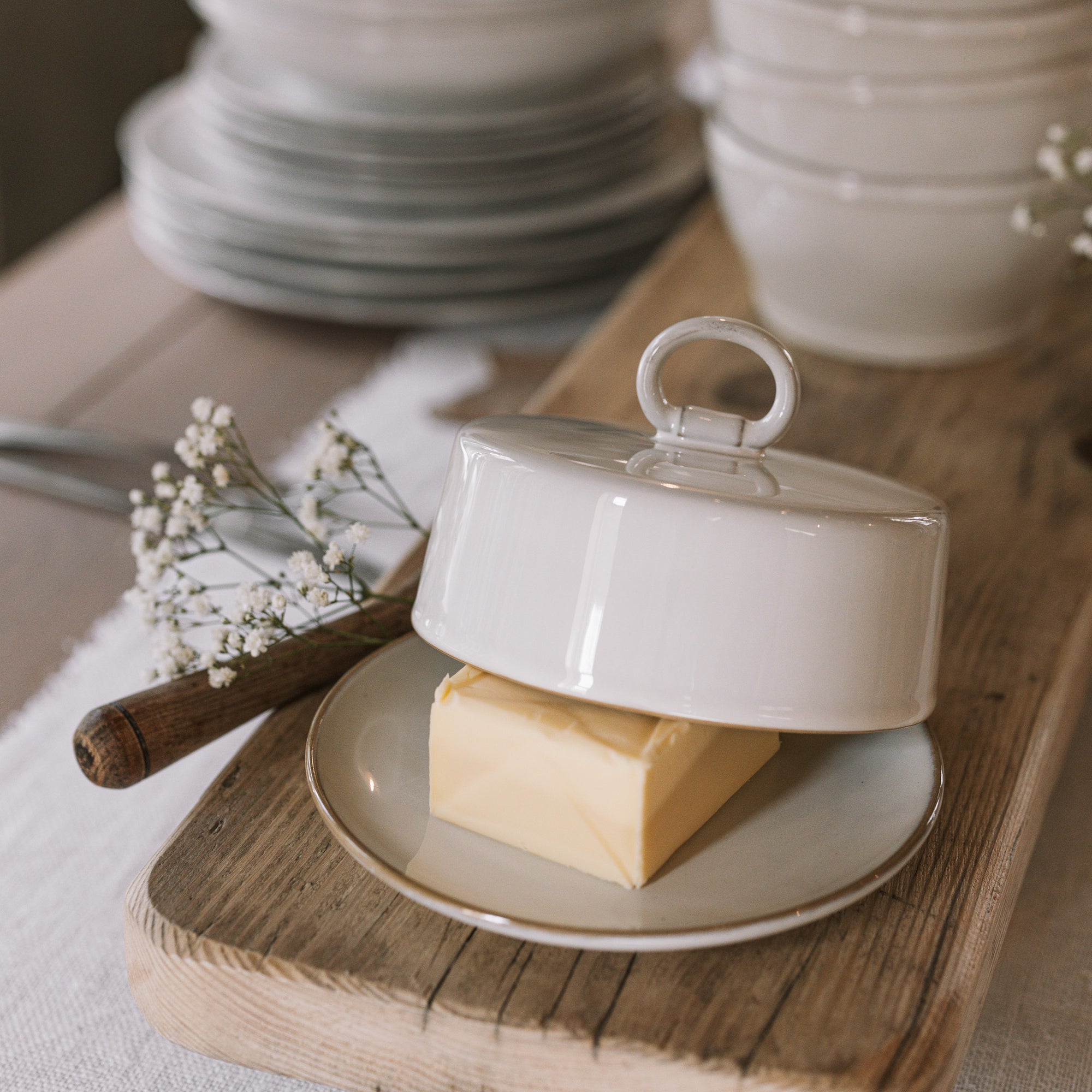 Off white butter dish with lid and loop to open on neutral wooden table with white table cloth.