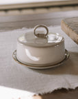 Off white butter dish with lid and loop to open on neutral wooden table with white table cloth.