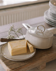 Off white butter dish with lid and loop to open on neutral wooden table with white table cloth.