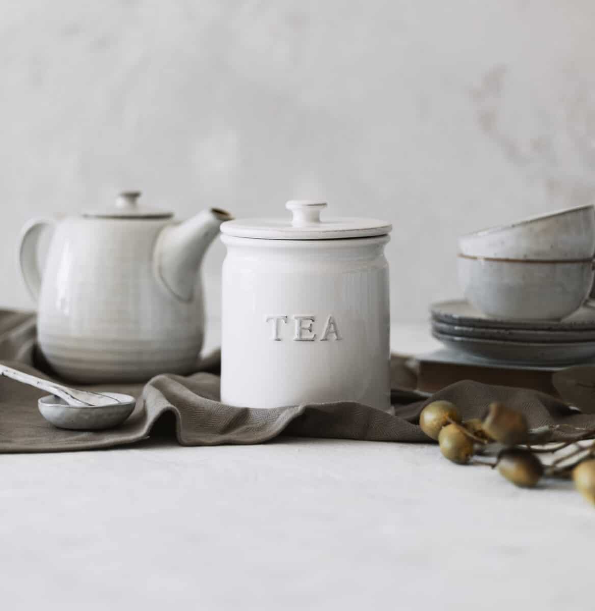 White ceramic tea storage jar with tea pot and crockery behind on tablecloth.
