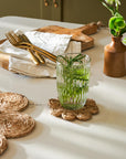 Glass of water with mint leaves on a woven jute coaster, surrounded by cutlery and a vase on a light surface.