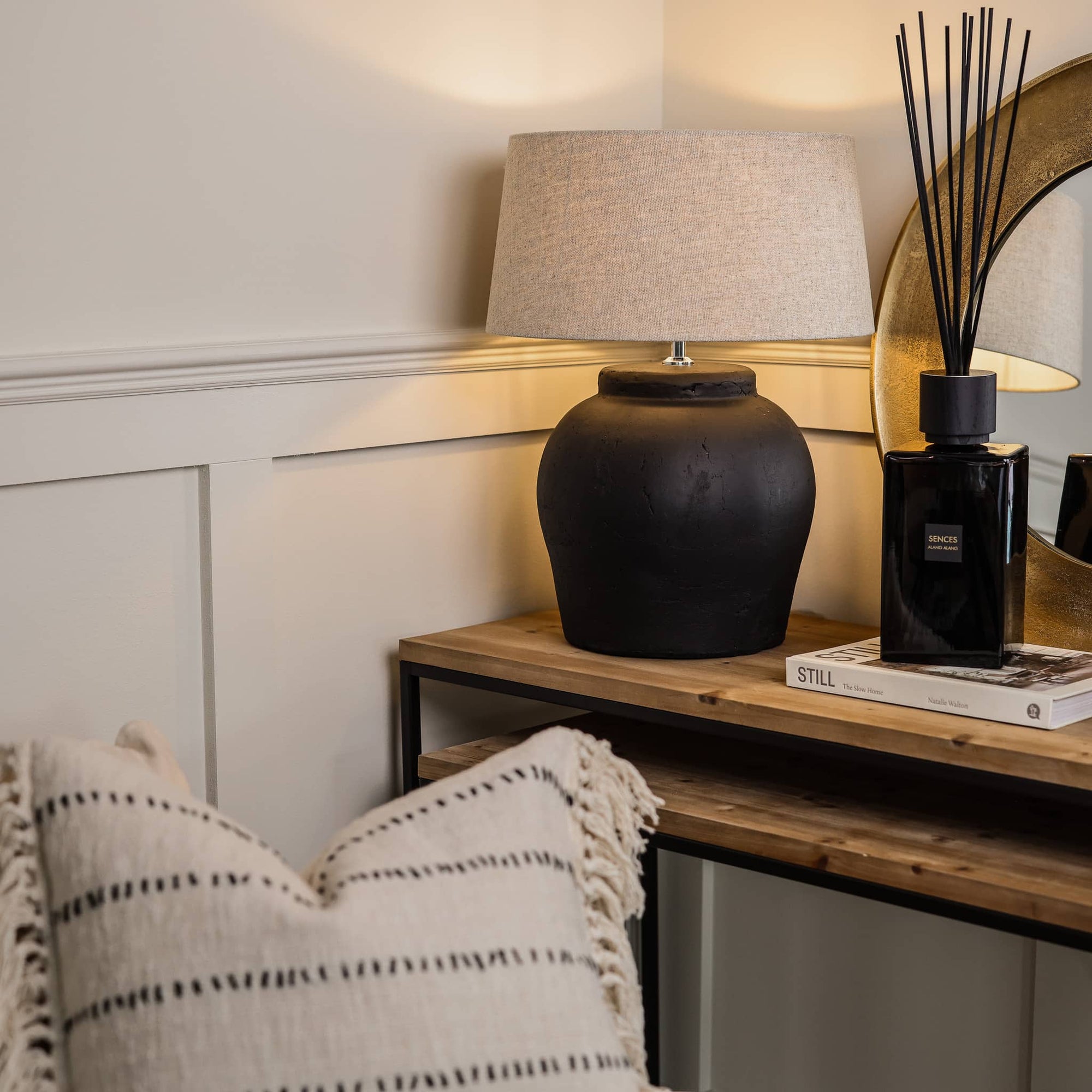 Matte black lamp with neutral shade, switched on on wooden console with black diffuser, white book and pillow in front.