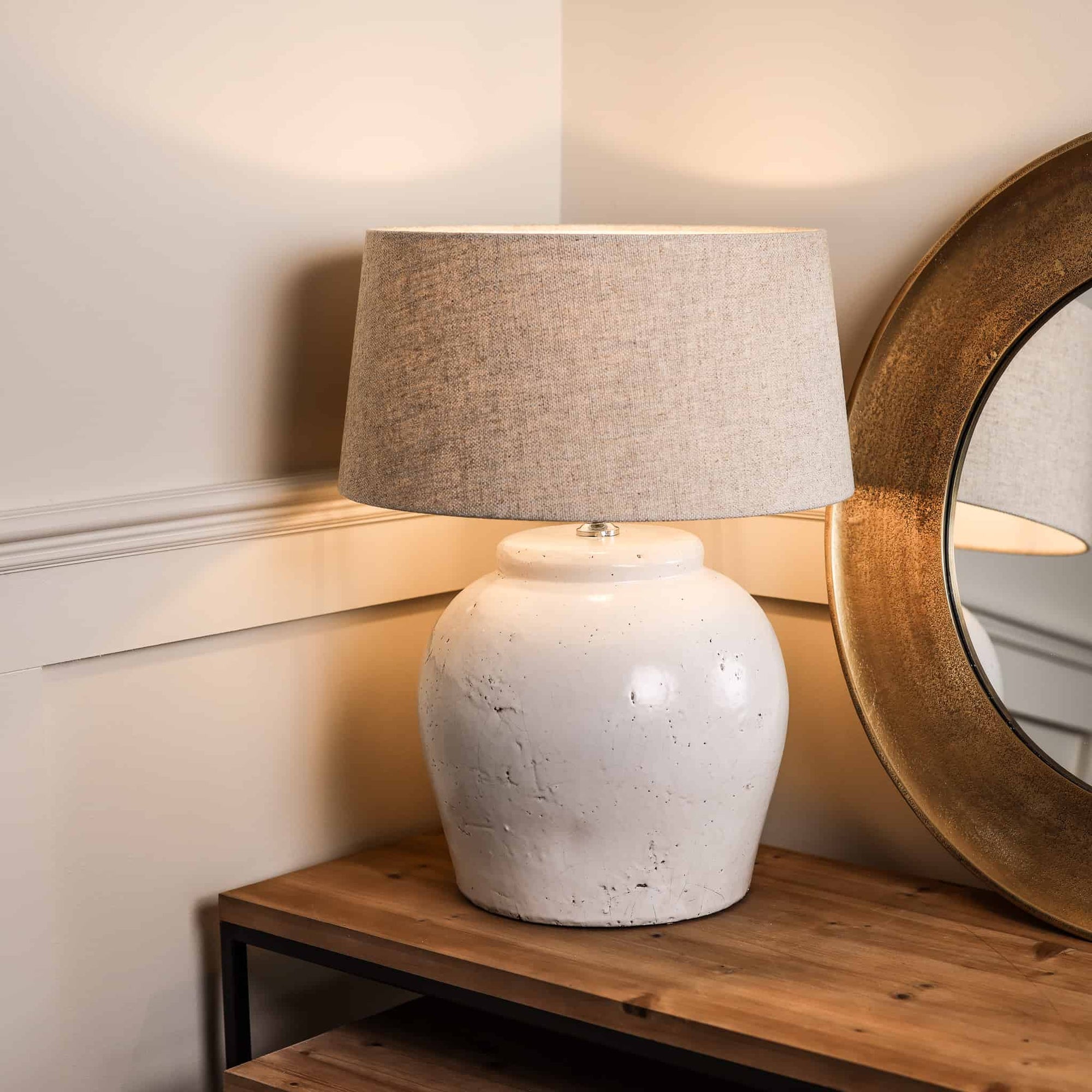 Distressed white lamp base with natural shade, switched on on wooden console.