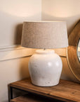 Distressed white lamp base with natural shade, switched on on wooden console.