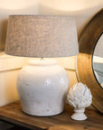Distressed white lamp base with natural shade, switched on on wooden console with acorn ornament.