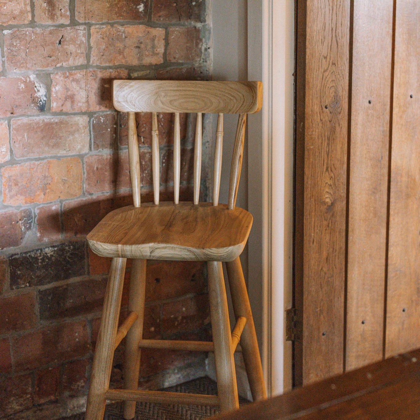 Wooden farmhouse bar stool with back against reclaimed brick wall.