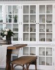 Large white armoire cabinet with glass doors filled with kitchen crockery, next to a dining table.