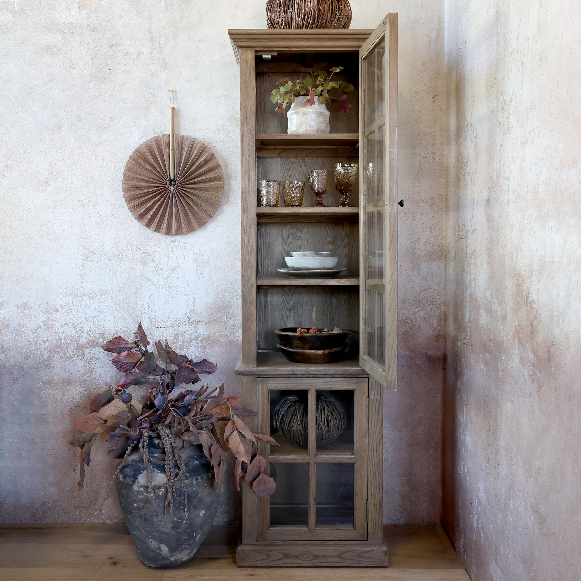 Natural wood display cabinet with glass door open, with dinnerware and glasses on shelves.