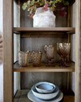 Close up of shelving on natural wood display cabinet, with amber glasses, a white vase and dinnerware.