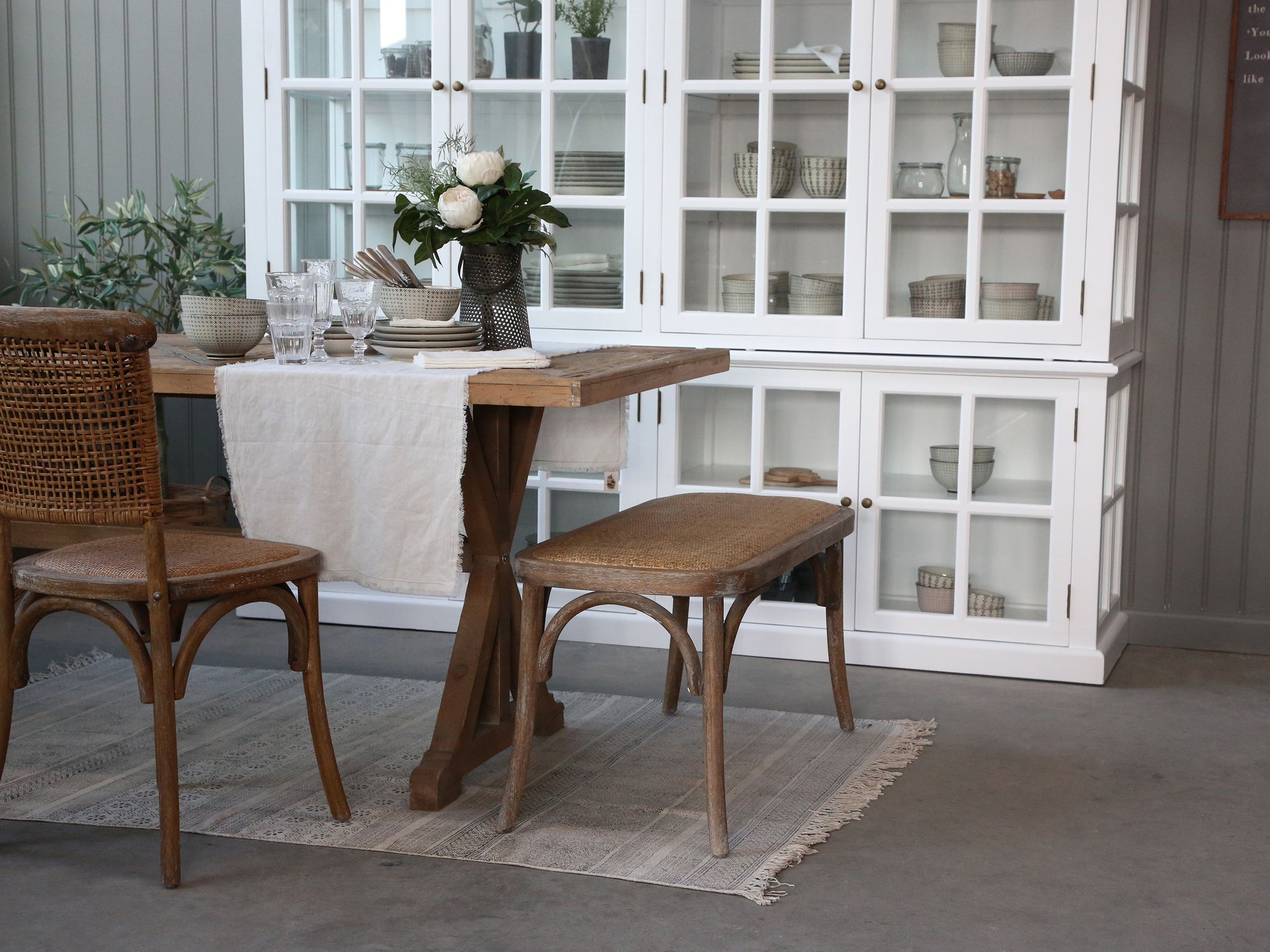 French Farmhouse Rattan Dining Bench at wooden dining table with white cupboard.
