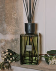 Green florist Sences reed diffuser on stone windowsill with flowers.