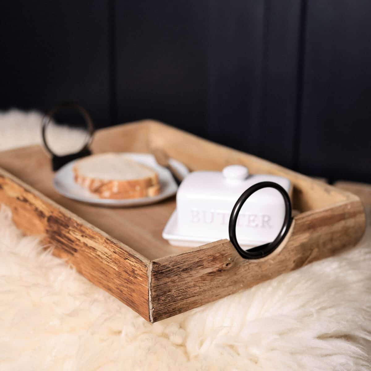 Wooden tray with black iron handles on a furry rug with butter and bread.