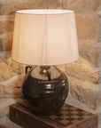 Large brown Table Lamp With Linen Shade on wooden chess board against stone wall.