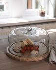 Recycled Glass Cake Dome filled with cheese, tomatoes and crackers with wooden base on wooden tale with gingham tea towel.