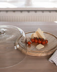 Recycled Glass Cake Dome filled with cheese, tomatoes and crackers with wooden base on wooden tale with gingham tea towel.