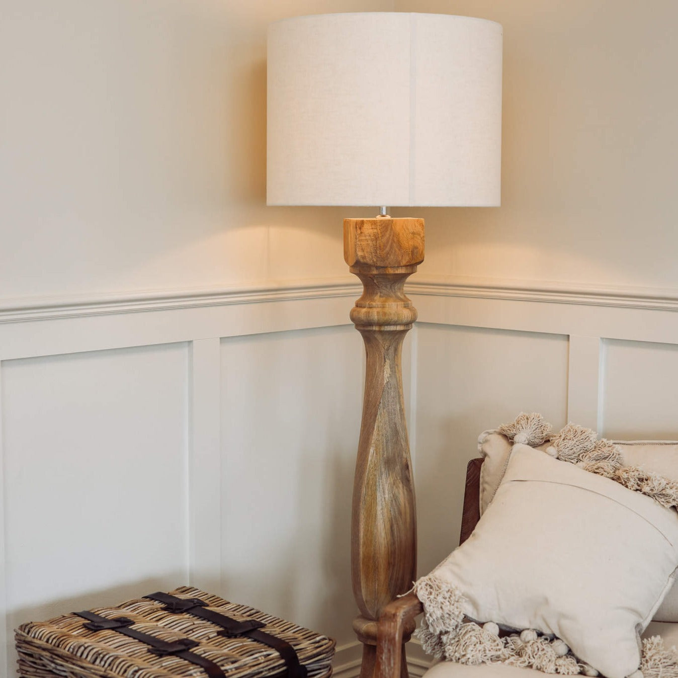 Tall wooden floor lamp switched on in the corner of a room with a chair and woven basket table.