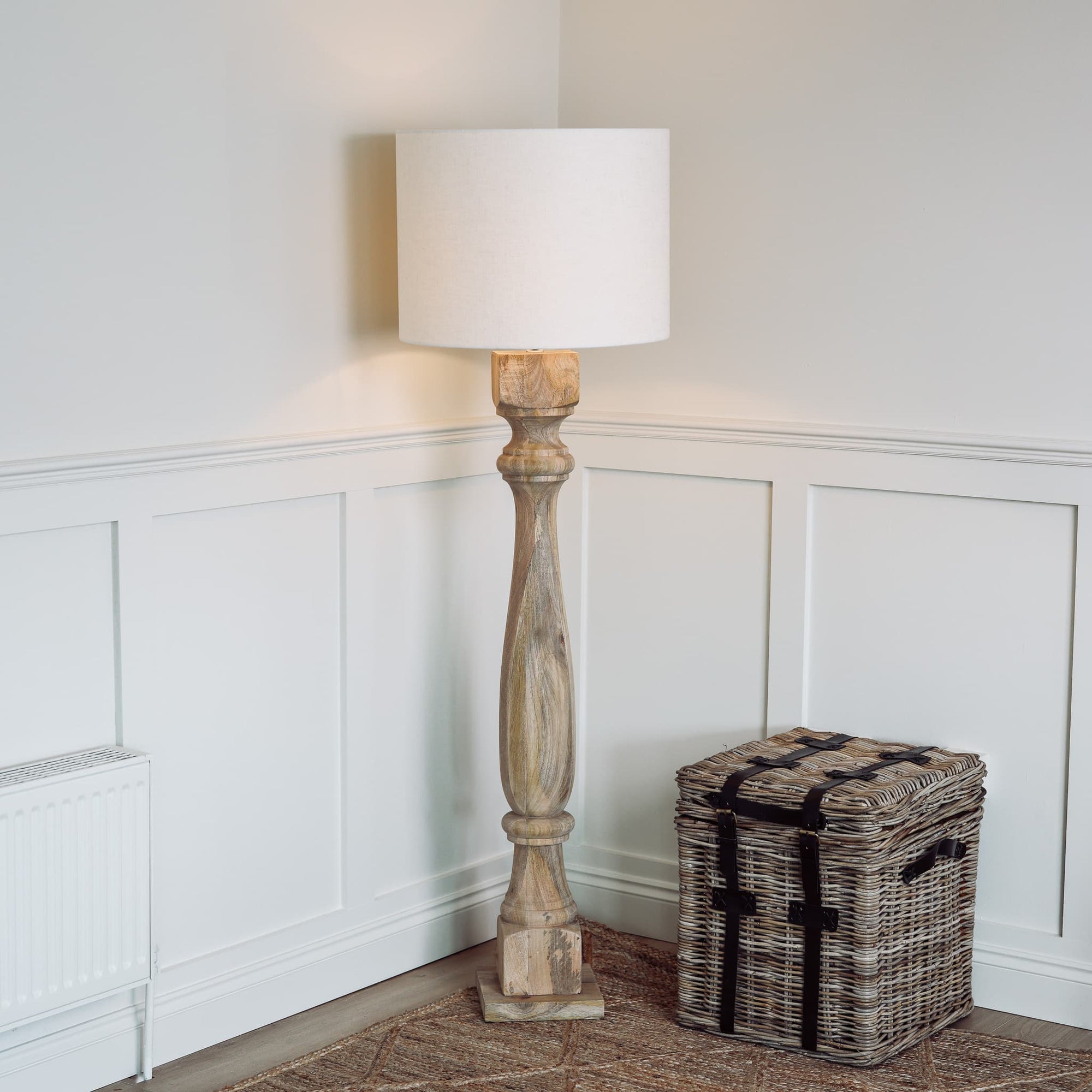 Tall wooden floor lamp switched on in the corner of a room, with a woven basket table.