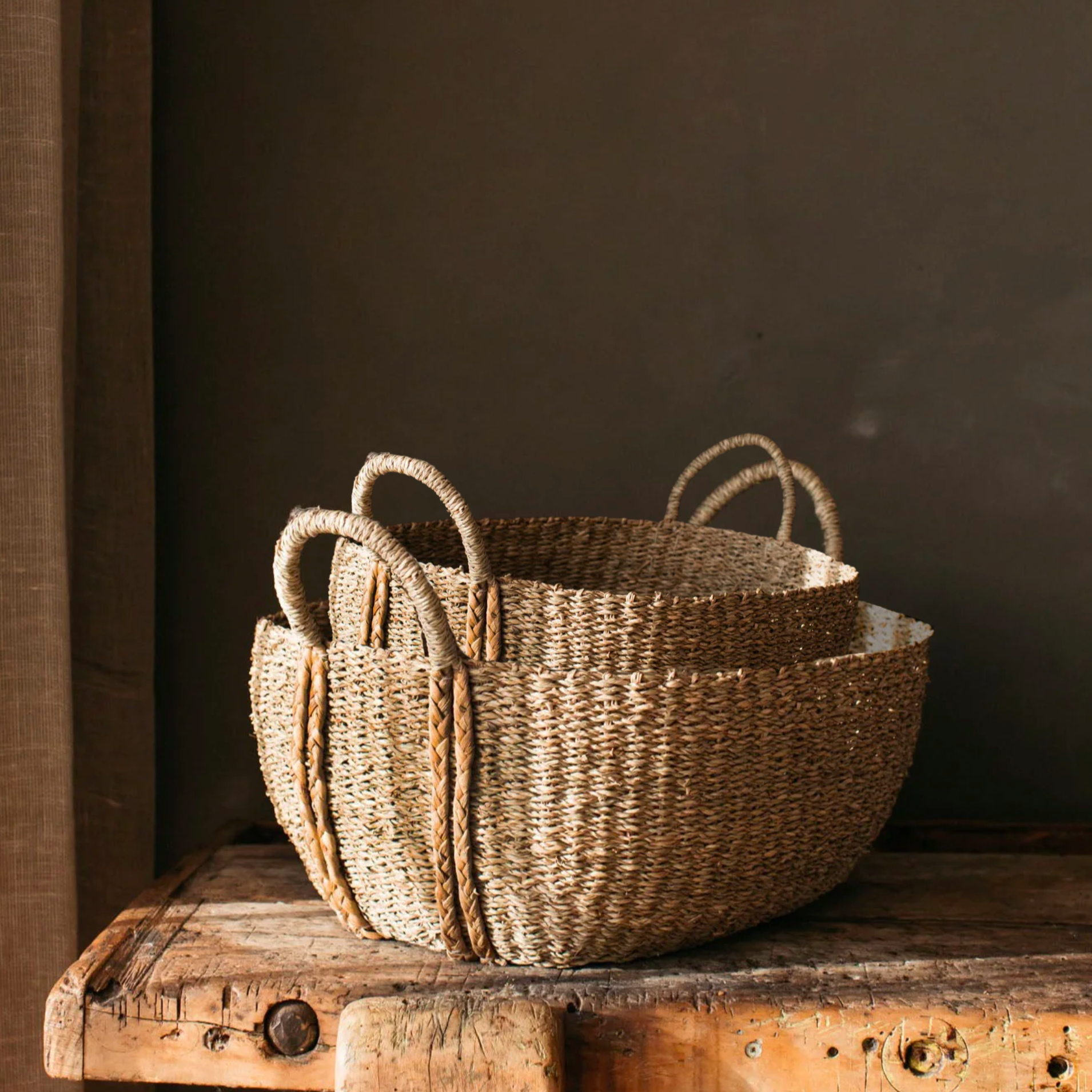 The Audrey Oval Seagrass Baskets - Set of Two placed on rustic wooden table against chocolate brown wall.