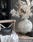 a dining table with a rustic vase with dried flowers, black crockery and a wooden dining chair.