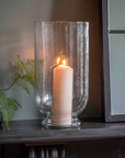 Glass hurricane vase filled with lit candle on a hearth.