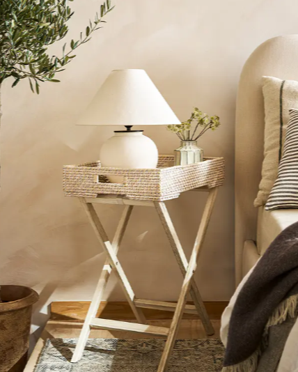 Rattan tray table next to bed with lamp and vase inside.