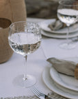 a textured wine glass filled with clear liquid on white linen tablecloth with lanterns in the background.