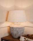 Medium grey distressed lamp, with white shade switched on. Styled with white mug and wooden tray.