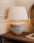 Medium grey distressed lamp with white shade switched on, on wooden console.