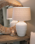 White ceramic table lamp with beaded detailing and linen lampshade on wooden table with small candle and mirror.