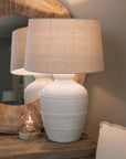 White ceramic table lamp with beaded detailing and linen lampshade on wooden table with small candle and mirror.