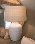 White ceramic table lamp with beaded detailing and linen lampshade on wooden table with small candle and mirror.